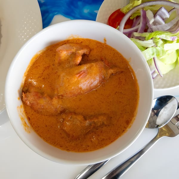 A white bowl filled with butter chicken, accompanied by a side salad and cutlery.