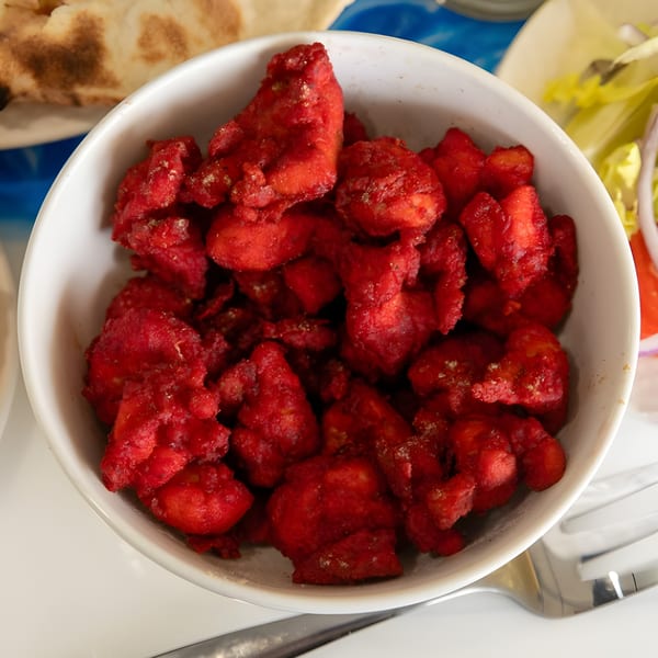 A white bowl filled with bright red chicken tikka, garnished with herbs, next to naan and salad.