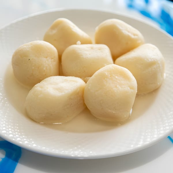 Seven light yellow Rasgulla, a traditional Indian sweet, soaked in syrup on a white plate.